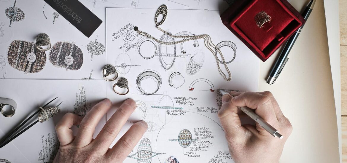 Hands sketching jewelry designs with tools and prototypes on a desk.